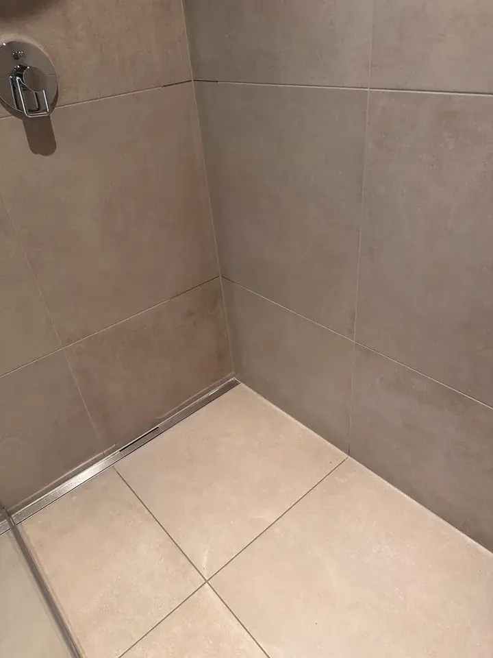 Duschecke mit grossen graubeigen Wandplatten, Armatur oben links, metallener Ablaufrinne an der Wand und hellen Bodenplatten