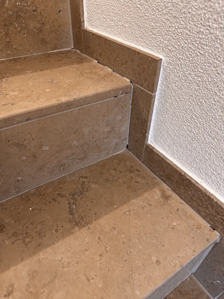 Braune Steinstufen mit Sockel an weisser, rauer Wand, in den Kanten zwischen Stufe und Sockel dunkle Linien