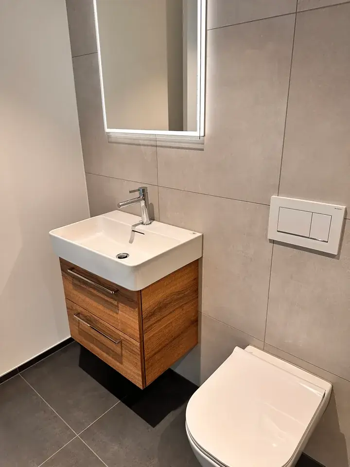 Bad mit Wand-WC, Waschtisch auf Holz-Unterschrank und beleuchtetem Spiegel vor grossformatigen beige-grauen Wandplatten