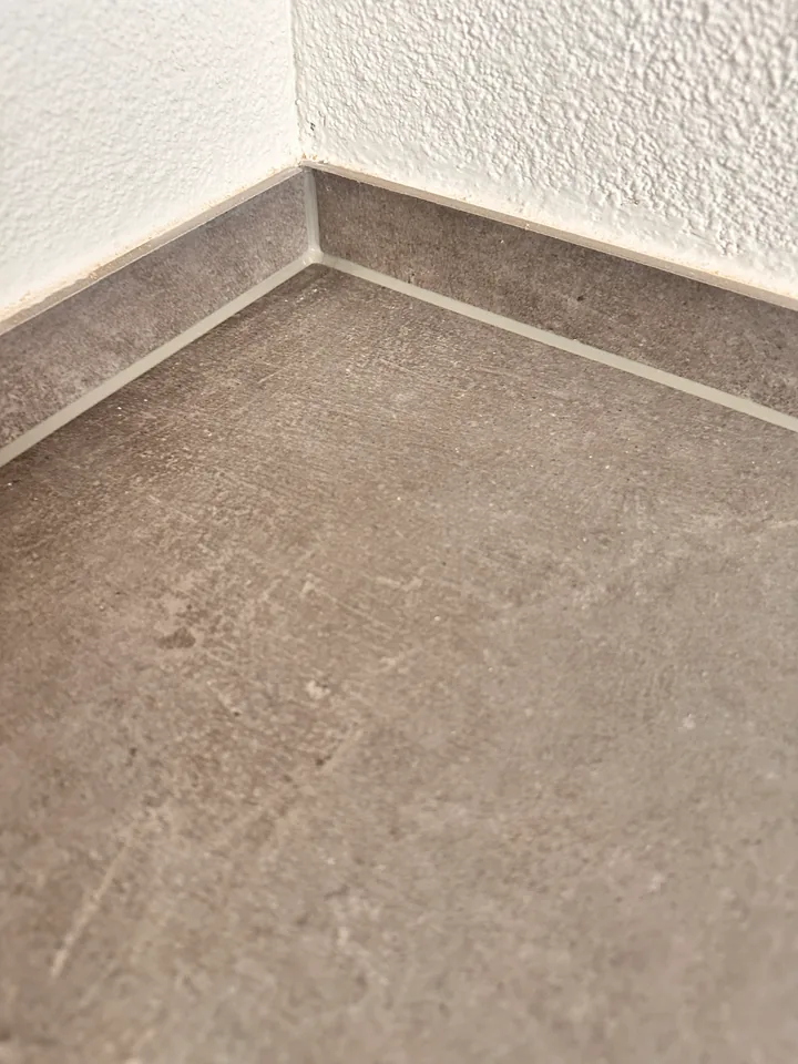 Graubrauner Plattenboden mit gleichfarbiger Sockelleiste und heller Fuge dazwischen vor rau verputzter weisser Wand